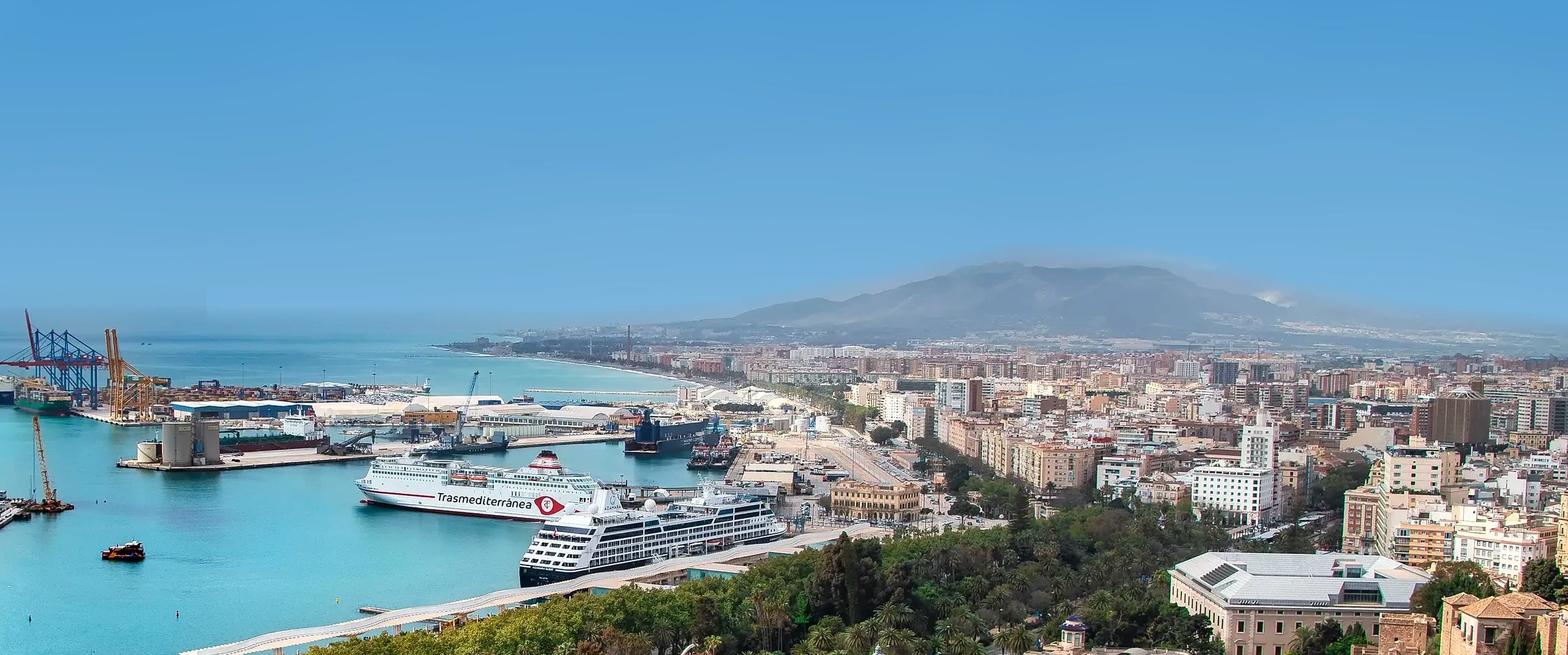 Vista aérea de Málaga de día, con cielo azul despejado, el puerto, la ciudad y las montañas al fondo.